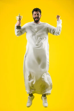 أنشطة ترفيهية ممتعة، النظر إلى الكاميرا بإيماءات الفرح والسعادة لفوز الفريق المفضل، تشجيع الأنديه والمنتخبات، رجل عربي خليجي اماراتي مبتسم يرتدي الكندورة يقفز عاليا ويمسك بيده الكأس الذهبي، رفع قبضة اليد بتعابير الانتصار والحماس، صورة الجسم بالكامل، خلفية صفراء