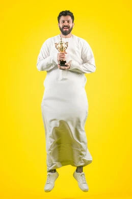 تشجيع الأنديه والمنتخبات، أنشطة ترفيهية ممتعة، النظر إلى الكاميرا بإيماءات الفرح والسعادة لفوز الفريق المفضل، رجل عربي خليجي اماراتي مبتسم يرتدي الكندورة يقفز عاليا ويمسك بيده الكأس الذهبي، تعابير الانتصار والحماس، صورة الجسم بالكامل، خلفية صفراء