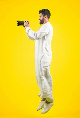 اتقان التصوير الفوتوغرافي باستخدام كاميرات رقمية عالية الدقة, مصور محترف يقوم بالتقاط صور تذكارية, ممارسة هواية التصوير بتعابير السرور, بورتريه من الجانب لشاب عربي خليجي اماراتي يرتدي الكندورة يمسك بيده كاميرا فوتوغرافية، صورة الجسم بالكامل، خلفية صفراء