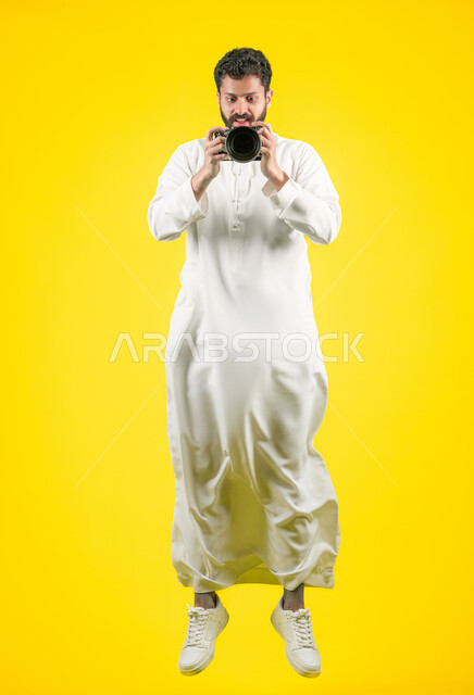 مصور محترف يقوم بالتقاط صور تذكارية, ممارسة هواية التصوير بتعابير السرور, اتقان التصوير الفوتوغرافي باستخدام كاميرات رقمية عالية الدقة, بورتريه لشاب عربي خليجي اماراتي يرتدي الكندورة يمسك بيده كاميرا فوتوغرافية، صورة الجسم بالكامل، خلفية صفراء