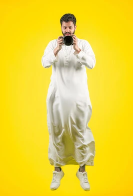 Professional photographer taking souvenir photos, practicing the hobby of photography with happy expressions, mastering photography using high-resolution digital cameras, portrait of a young Emirati Gulf Arab man wearing a kandura holding a camera, full body photo, yellow background