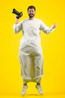 ممارسة هواية التصوير بتعابير السرور, اتقان التصوير الفوتوغرافي باستخدام كاميرات رقمية عالية الدقة, مصور محترف يقوم بالتقاط صور تذكارية, بورتريه لشاب عربي خليجي اماراتي يرتدي الكندورة يقفز عاليا ويمسك بيده كاميرا فوتوغرافية، صورة الجسم بالكامل، خلفية صفراء