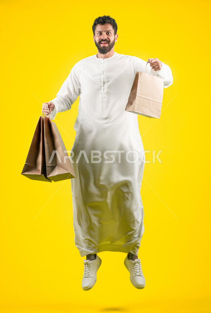 المشتريات التجارية والهدايا, القفز عاليا والنظر إلى الكاميرا بتعابير السرور، التبضع وأخذ الاحتياجات اللازمة, مواسم العروض والتخفيضات, الاستفادة من الحسومات والتصفيات، رجل عربي خليجي اماراتي يرتدي الكندورة و يمسك بيده أكياس ورقية، صورة الجسم بالكامل، خلفية صفراء