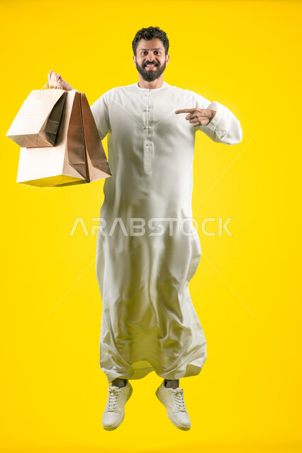 The joy of shopping, taking advantage of sales and clearance seasons, pointing with the index finger at commercial purchases and gifts, shopping and taking the necessary needs, jumping high with happiness, portrait of an Arab Gulf Emirati man wearing a kandura and holding paper bags in his hand, full body image, yellow background