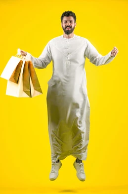 الاستفادة من الحسومات والتصفيات، القفز عاليا والنظر إلى الكاميرا بتعابير السرور، المشتريات التجارية والهدايا, التبضع وأخذ الاحتياجات اللازمة, مواسم العروض والتخفيضات, رجل عربي خليجي اماراتي يرتدي الكندورة و يمسك بيده أكياس ورقية، صورة الجسم بالكامل، خلفية صفراء