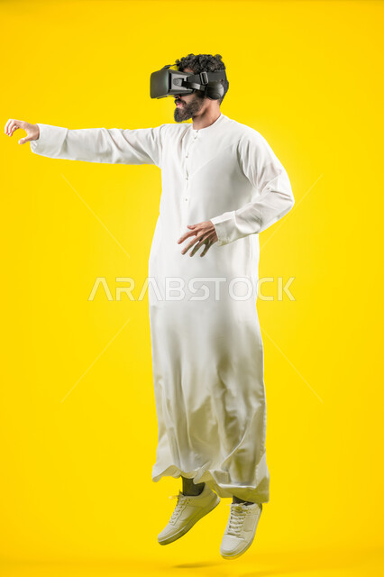 القفز عاليا ورفع اليدين لمحاولة امساك شيء ما، المتعة بمشاهدة الأفلام ومحاكاة العالم الخيالي, بورتريه لشاب عربي خليجي اماراتي يرتدي الكندورة ونظارة في آر ثلاثية الأبعاد, التفاعل والاندماج مع ألعاب الواقع الافتراضي, الجسم بالطول الكامل، خلفية صفراء