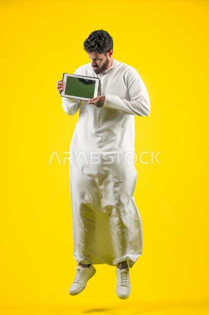 Using modern and advanced technology, completing work tasks remotely via a portable tablet, displaying a blank black screen via the tablet, portrait of an Arab Gulf Emirati man wearing a kandura, full body length, yellow background