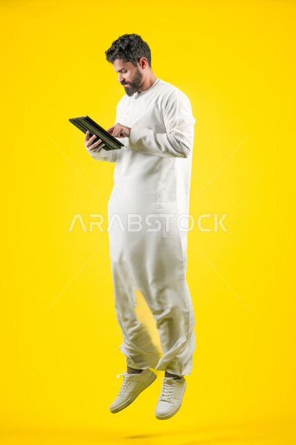 استخدام الجهاز اللوحي في إنجاز مهام العمل، شاب سعودي يتصفح تطبيقات التواصل الاجتماعي، استعمال التكنولوجيا الحديثة والمتطورة، بورتريه لرجل عربي خليجي اماراتي يرتدي الزي الكاجوال يستخدم جهاز التابلت بإيماءات التركيز، طول الجسم بالكامل، خلفية صفراء