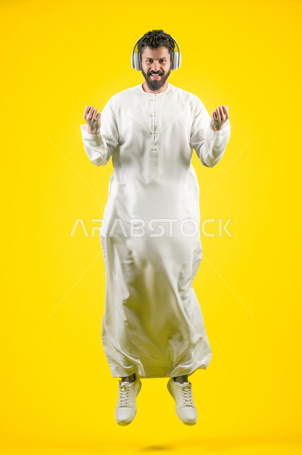تسجيل مقطع صوتي غنائي، مواهب غنائية فنية، بورتريه لرجل عربي خليجي اماراتي يرتدي الكندورة يضع الهيدفون، إيماءات السعادة و الاندماج مع الأغنية، الاستماع إلى الموسيقى, صورة الجسم بالكامل، خلفيه صفراء