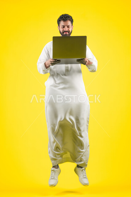استخدام اللابتوب في إنجاز مهام العمل عن بعد، القفز عاليا بسعادة، استعمال التكنولوجيا الحديثة والمتطورة، بورتريه لرجل عربي خليجي اماراتي يرتدي الكندورة يستخدم جهاز الحاسوب بإيماءات التركيز، طول الجسم بالكامل، خلفية صفراء