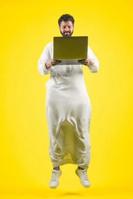 استخدام اللابتوب في إنجاز مهام العمل عن بعد، القفز عاليا بسعادة، استعمال التكنولوجيا الحديثة والمتطورة، بورتريه لرجل عربي خليجي اماراتي يرتدي الكندورة يستخدم جهاز الحاسوب بإيماءات التركيز، طول الجسم بالكامل، خلفية صفراء