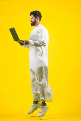استعمال التكنولوجيا الحديثة والمتطورة، استخدام اللابتوب في إنجاز مهام العمل عن بعد، القفز عاليا بسعادة، بورتريه من الجانب لرجل عربي خليجي اماراتي يرتدي الكندورة يستخدم جهاز الحاسوب بإيماءات التركيز، طول الجسم بالكامل، خلفية صفراء