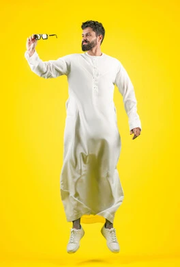 اقتناء نظارات جديدة، الأناقة والوسامة والاهتمام بالمظهر الخارجي، استخدام العدسات الطبية الملونة لحماية العين من أشعة الشمس، بورتريه لرجل عربي خليجي اماراتي  يرتدي الكندورة يحمل نظارة شمسية سوداء، القفز عاليا بتعابير السعادة، الجسم بالطول الكامل, خلفية صفراء