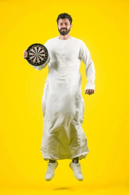 The concept of focus and goal setting, entertainment and sports skills, looking at the camera and jumping high with expressions of pleasure and happiness, portrait of a smiling Emirati Gulf Arab man wearing a kandura holding a magnetic darts game, full body, yellow background