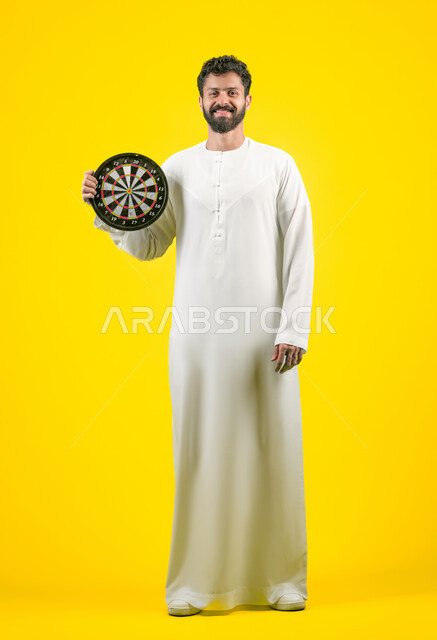 النظر الى الكاميرا بتعابير السرور والسعادة, مفهوم التركيز وتحديد الأهداف, تسليه وترفيه و مهارات رياضية، بورتريه لرجل عربي خليجي اماراتي مبتسم يرتدي الكندورة يحمل لعبة السهام المغناطيسية، الجسم بالطول الكامل، خلفية صفراء