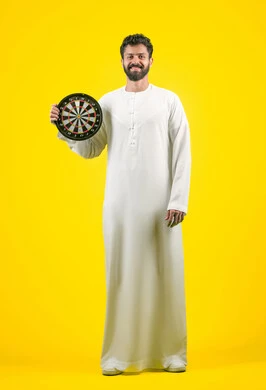 النظر الى الكاميرا بتعابير السرور والسعادة, مفهوم التركيز وتحديد الأهداف, تسليه وترفيه و مهارات رياضية، بورتريه لرجل عربي خليجي اماراتي مبتسم يرتدي الكندورة يحمل لعبة السهام المغناطيسية، الجسم بالطول الكامل، خلفية صفراء