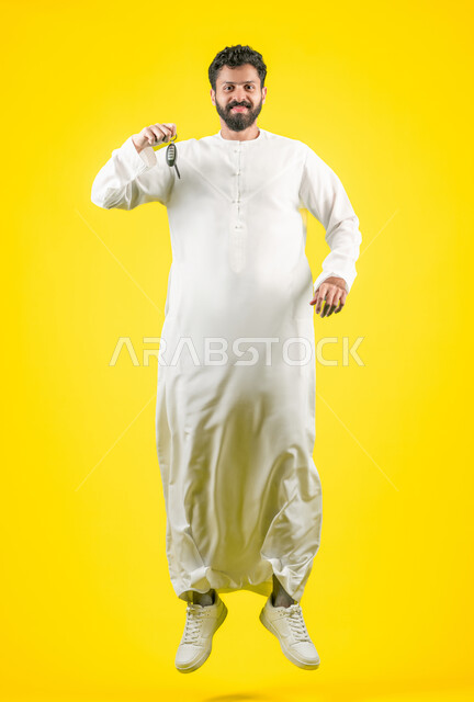الحصول على رخصة القيادة، استلام وتسليم المفاتيح، القفز عاليا بإيماءات الحماس لامتلاك سيارة جديدة، خدمه صف السيارات بالمواقف الخاصه, بورتريه لشاب عربي خليجي اماراتي يرتدي الكندورة يمسك بيده مفتاح التحكم عن بعد, الجسم بالطول الكامل، خلفية صفراء