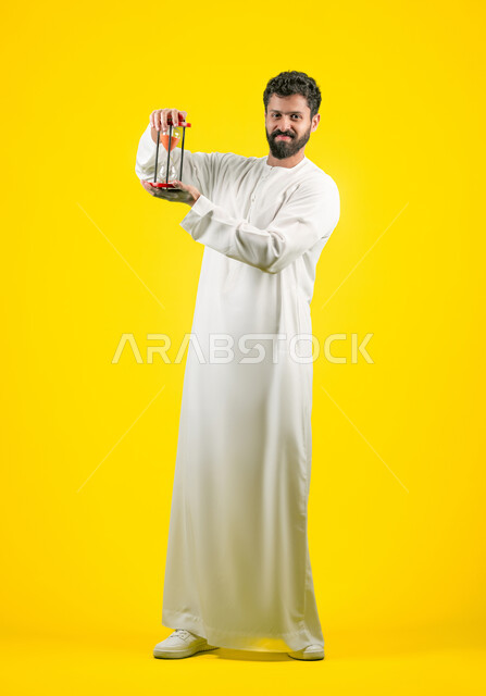 Business management and schedule organization concept, portrait of a young Emirati Gulf Arab man wearing a kandura holding a glass sand hourglass (bankam) in his hand, looking at the camera with gestures of joy and pleasure, monitoring the timing of the sand, realizing the value of precious moments, full body, yellow background