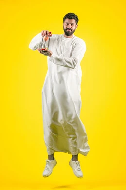 Realizing the value of precious moments, the concept of business management and schedule organization, portrait of a young Emirati Gulf Arab man wearing a kandura holding a glass sand hourglass (bankam) in his hand, jumping in the air with gestures of joy and happiness, performing sand timing, full body, yellow background