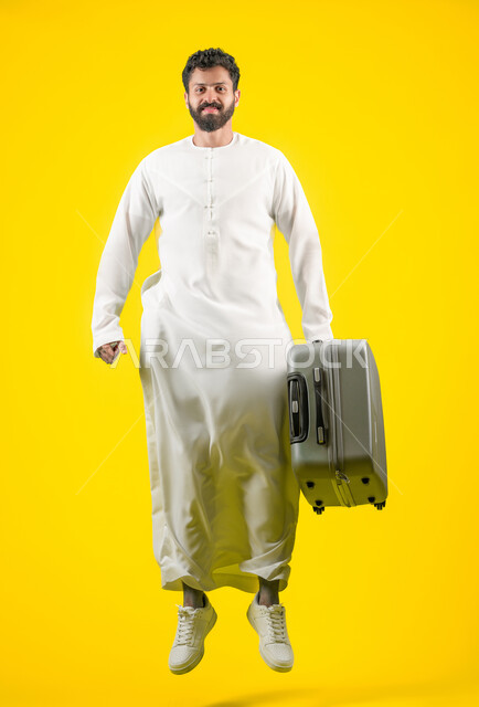 القفز عاليا والنظر إلى الكاميرا بايماءات السعادة والفرح، القيام بجولات ترفيهية في إجازة الصيف, بورتريه لشاب عربي خليجي اماراتي يرتدي الكندورة يحمل حقيبة السفر, مفهوم السياحة والسفر، الاستعداد لقضاء رحلة صيفية ممتعة, صورة الجسم بالطول الكامل، خلفية صفراء
