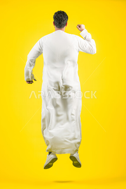 Cheering for the victory of the favorite team, increasing muscle endurance, mastering the art of boxing, portrait from the back of an Arab Gulf Emirati young man wearing a kandura, jumping high and raising his fists with gestures of strength, practicing self-defense, full body, yellow background