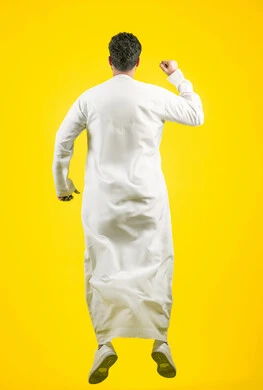 تشجيع وفوز الفريق المفضل، زيادة قدرة العضلات على التحمل، إتقان فنون الملاكمة، بورتريه من الخلف لشاب عربي خليجي اماراتي يرتدي الكندورة يقفز عاليا ويرفع قبضة يديه بإيماءات القوة، ممارسة رياضة الدفاع عن النفس، الجسم بالطول الكامل، خلفية صفراء