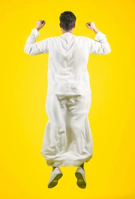زيادة قدرة العضلات على التحمل، إتقان فنون الملاكمة، تشجيع وفوز الفريق المفضل، بورتريه من الخلف لشاب عربي خليجي اماراتي يرتدي الكندورة يقفز عاليا ويرفع قبضات يديه بإيماءات القوة، ممارسة رياضة الدفاع عن النفس، الجسم بالطول الكامل، خلفية صفراء