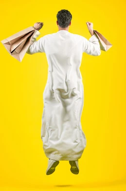 المتعة في التسوق، الاستفادة من مواسم العروض والحسومات والتصفيات، المشتريات التجارية والهدايا, التبضع وأخذ الاحتياجات اللازمة, القفز عاليا بسعادة، بورتريه من الخلف لرجل عربي خليجي اماراتي يرتدي الكندورة و يمسك بيده أكياس ورقية، صورة الجسم بالكامل، خلفية صفراء