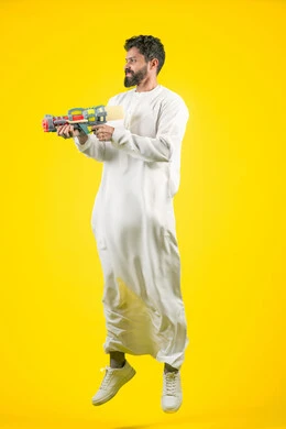 Having fun with water games in the summer, portrait of a young Emirati Gulf Arab man wearing a kandura holding a plastic water gun playing with gestures of pleasure and happiness, jumping high with expressions of enthusiasm and joy, the concept of entertainment and amusement, full body, braided background