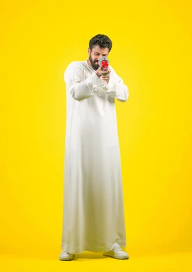 مفهوم الترفيه عن النفس والتسلية، قضاء وقت ممتع بالألعاب المائية في فصل الصيف، بورتريه لشاب عربي خليجي اماراتي يرتدي الكندورة يحمل مسدس مائي بلاستيكي يقوم باللعب بإيماءات السرور والسعادة، الجسم بالطول الكامل، خلفية ضفراء