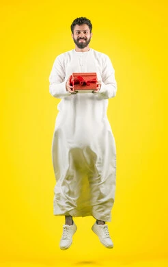 Expressing love, appreciation and respect, giving gifts on holidays and special occasions, the concept of thanks and gratitude, jumping high and looking at the camera with gestures of joy and happiness, portrait of a young Emirati Arab man wearing a kandura holding a gift box, full body, yellow background