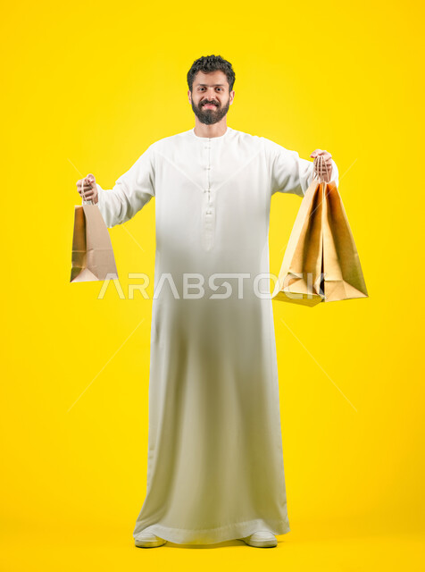 الاستفادة من الحسومات والتصفيات، النظر إلى الكاميرا بتعابير السرور، المشتريات التجارية والهدايا, التبضع وأخذ الاحتياجات اللازمة, مواسم العروض والتخفيضات, رجل عربي خليجي اماراتي يرتدي الكندورة و يمسك بيده أكياس ورقية، صورة الجسم بالكامل، خلفية صفراء