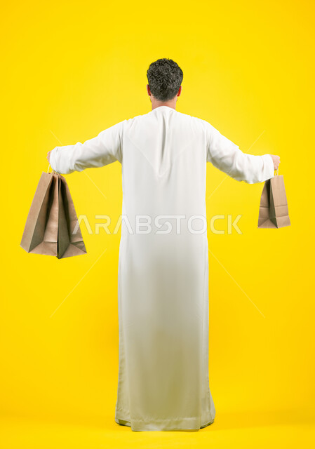 مواسم العروض والتخفيضات, الاستفادة من الحسومات والتصفيات، المشتريات التجارية والهدايا, التبضع وأخذ الاحتياجات اللازمة, بورتريه من الخلف لرجل عربي خليجي اماراتي يرتدي الكندورة و يمسك بيده أكياس ورقية، صورة الجسم بالكامل، خلفية صفراء