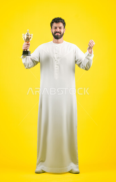 Fun recreational activities, looking at the camera with gestures of joy and happiness for the victory of the favorite team, cheering for clubs and national teams, a smiling Emirati Gulf Arab man wearing a kandura holding the golden cup in his hand, raising his fist with expressions of victory and enthusiasm, full body image, yellow background
