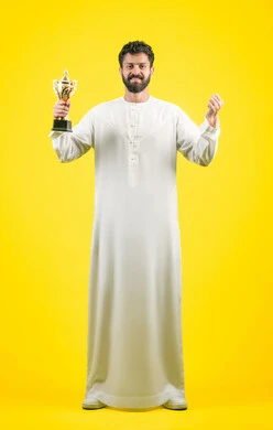 أنشطة ترفيهية ممتعة، النظر إلى الكاميرا بإيماءات الفرح والسعادة لفوز الفريق المفضل، تشجيع الأنديه والمنتخبات، رجل عربي خليجي اماراتي مبتسم يرتدي الكندورة يمسك بيده الكأس الذهبي، رفع قبضة اليد بتعابير الانتصار والحماس، صورة الجسم بالكامل، خلفية صفراء