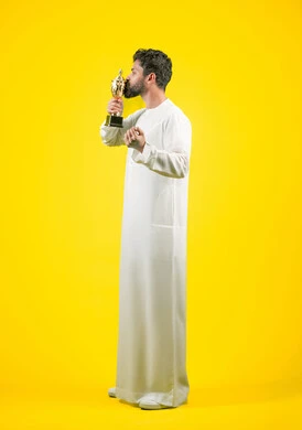 رفع قبضة اليد بتعابير الانتصار والحماس، أنشطة ترفيهية ممتعة، إيماءات الفرح والسعادة لفوز الفريق المفضل، تشجيع الأنديه والمنتخبات، رجل عربي خليجي اماراتي مبتسم يرتدي الكندورة يقوم بتقبيل الكأس الذهبي، صورة الجسم بالكامل، خلفية صفراء