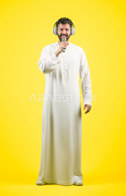 تسجيل مقطع صوتي غنائي، مواهب غنائية فنية، إيماءات السعادة و الاندماج مع الأغنية، بورتريه لرجل عربي خليجي اماراتي يرتدي الكندورة يضع سماعة الرأس ويقوم بالغناء باستخدام الميكروفون بتعابير الحماس, صورة الجسم بالكامل، خلفيه صفراء