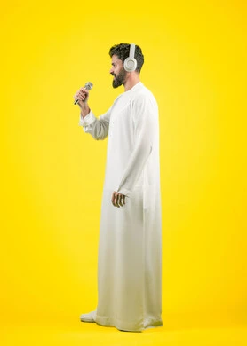 مواهب غنائية فنية، تسجيل مقطع صوتي غنائي، إيماءات السعادة و الاندماج مع الأغنية، بورتريه من الجانب لشاب عربي خليجي اماراتي يرتدي الكندورة يضع سماعة الرأس ويقوم بالغناء باستخدام الميكروفون بتعابير الحماس, صورة الجسم بالكامل، خلفيه صفراء