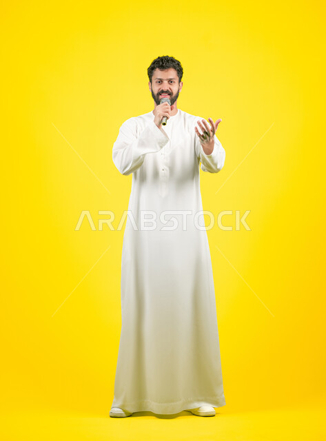 الاندماج مع الأغنية، مواهب غنائية فنية، النظر إلى الكاميرا بإيماءات السرور، تسجيل مقطع صوتي غنائي، بورتريه لشاب عربي خليجي اماراتي يرتدي الكندورة يقوم بالغناء باستخدام الميكروفون بتعابير الحماس, صورة الجسم بالكامل، خلفيه صفراء