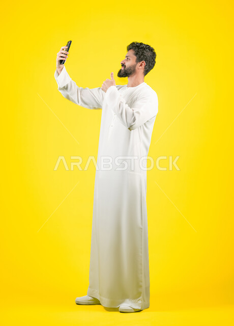 Live broadcast on social media, thumbs up gesture, portrait of an Emirati Gulf Arab man wearing a kandura taking a souvenir photo with a mobile phone, answering a video call, full body photo, yellow background