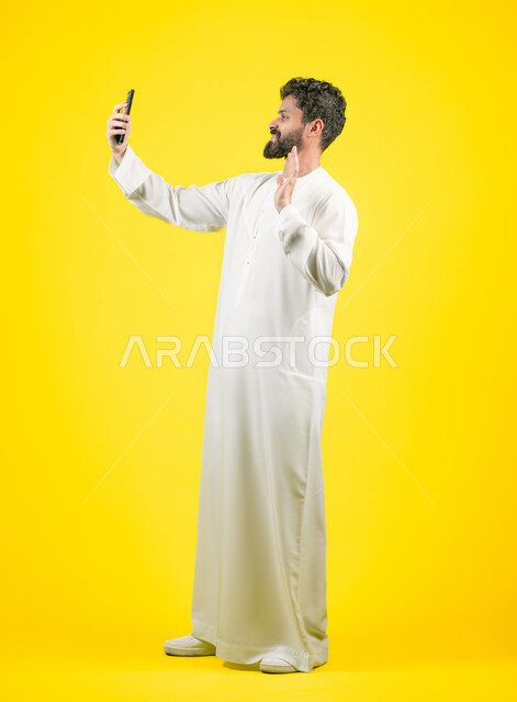 رفع اليد للأعلى بايماءات القاء التحية والسلام، إجراء بث مباشر على إحدى وسائل التواصل الاجتماعي, بوتريه لرجل عربي خليجي اماراتي يرتدي الكندورة يقوم بالتقاط صور تذكارية باستخدام الهاتف الجوال, الرد على مكالمة فيديو هاتفية، صورة الجسم بالكامل، خلفية صفراء