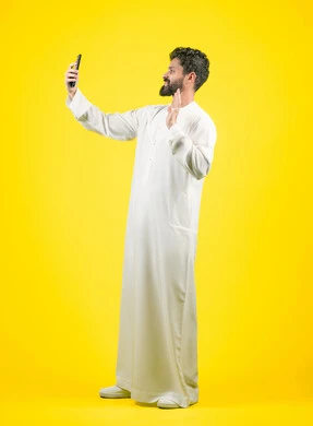 Raising hand up in greeting gesture, live broadcast on social media, portrait of an Emirati Gulf Arab man wearing a kandura taking a souvenir photo using a mobile phone, answering a video call, full body photo, yellow background