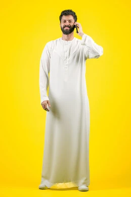 التواصل مع الأهل والأصدقاء, النظر إلى الكاميرا بإيماءات البشاشة، متابعة آخر الأخبار والمستجدات حول العالم, دمج التقنيات الحديثة والتكنولوجيا بالحياة العملية, بورتريه لشاب عربي خليجي اماراتي يرتدي الكندورة يجري مكالمة هاتفية، الجسم بالطول الكامل، خلفية صفراء