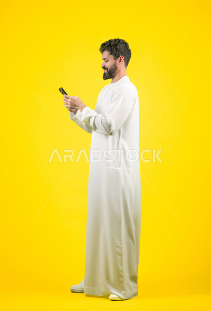 دمج التقنيات الحديثة والتكنولوجيا بالحياة العملية, التواصل مع الأهل والأصدقاء, متابعة آخر الأخبار والمستجدات حول العالم, بورتريه من الجانب لشاب عربي خليجي اماراتي يرتدي الكندورة يقوم باستخدام الهاتف المحمول، الجسم بالطول الكامل، خلفية صفراء