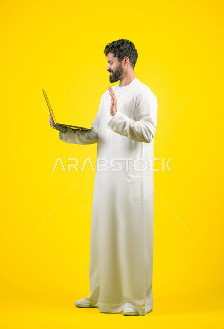 اجراء اجتماع عمل اونلاين، استعمال التكنولوجيا الحديثة والمتطورة، استخدام اللابتوب في إنجاز مهام العمل عن بعد، بورتريه لرجل عربي خليجي اماراتي يرتدي الكندورة يستخدم جهاز الحاسوب بإيماءات التركيز، طول الجسم بالكامل، خلفية صفراء
