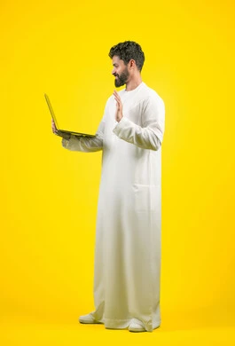 اجراء اجتماع عمل اونلاين، استعمال التكنولوجيا الحديثة والمتطورة، استخدام اللابتوب في إنجاز مهام العمل عن بعد، بورتريه لرجل عربي خليجي اماراتي يرتدي الكندورة يستخدم جهاز الحاسوب بإيماءات التركيز، طول الجسم بالكامل، خلفية صفراء