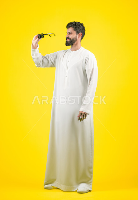اقتناء نظارات جديدة، الأناقة والوسامة والاهتمام بالمظهر الخارجي، استخدام العدسات الطبية الملونة لحماية العين من أشعة الشمس، بورتريه لرجل عربي خليجي اماراتي  يرتدي الكندورة يحمل نظارة شمسية سوداء بتعابير السعادة، الجسم بالطول الكامل, خلفية صفراء