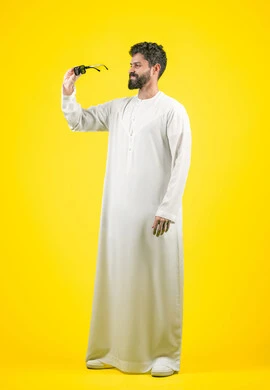 Buying new glasses, elegance, handsomeness and interest in outward appearance, using colored medical lenses to protect the eyes from the sun's rays, portrait of an Arab Gulf Emirati man wearing a kandura holding black sunglasses with a happy expression, full body, yellow background