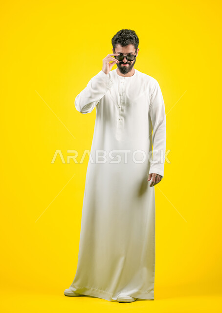 الأناقة والوسامة والاهتمام بالمظهر الخارجي، استخدام العدسات الطبية الملونة لحماية العين من أشعة الشمس، بورتريه لرجل عربي خليجي اماراتي  يرتدي الكندورة ينظر إلى الكاميرا بإيماءات الثقة بالنفس ويضع نظارة شمسية سوداء، الجسم بالطول الكامل, خلفية صفراء