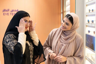 مفهوم الموضة والاناقة والاهتمام بالمظهر الخارجي، امرأة اماراتية تساعد صديقتها فى اختيار النظاره الملائمه لملامح الوجه ، التأثير الإيجابي للتسوق على المرأة، اختيار نظارة شمسيه مناسبة ،متعة الشراء من احدي المولات الاماراتية، صديقتان عربيتان خليجيتان سعوديتان ترتديان العباءة احداهما تقوم بقياس النظارة